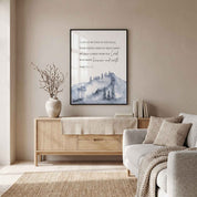 Framed Psalm 121:1–2 mountain wall art in blue gray watercolor, displayed above a light wood sideboard with books and a vase in a living room.