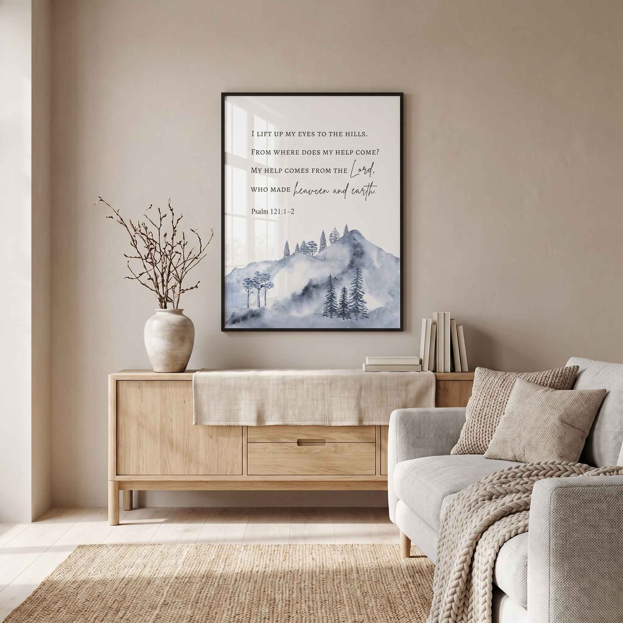 Framed Psalm 121:1–2 mountain wall art in blue gray watercolor, displayed above a light wood sideboard with books and a vase in a living room.