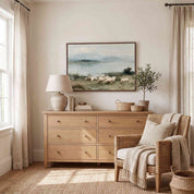 Framed Psalm 23 vintage landscape canvas art above a light wood dresser with a wicker armchair in a serene warm-toned room, depicting sheep grazing by calm waters.