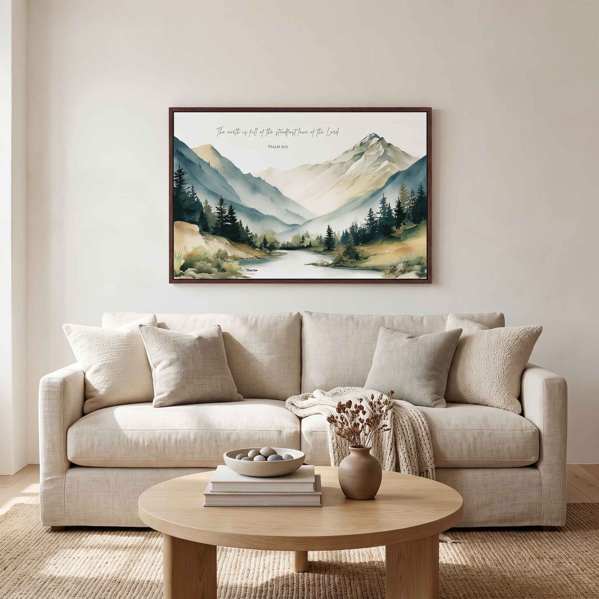 Framed vintage Christian mountain landscape wall art titled Psalm 33:5 featuring mountains, pine trees, and a river, displayed on a light beige wall above a beige sofa in a soft organic modern living room.