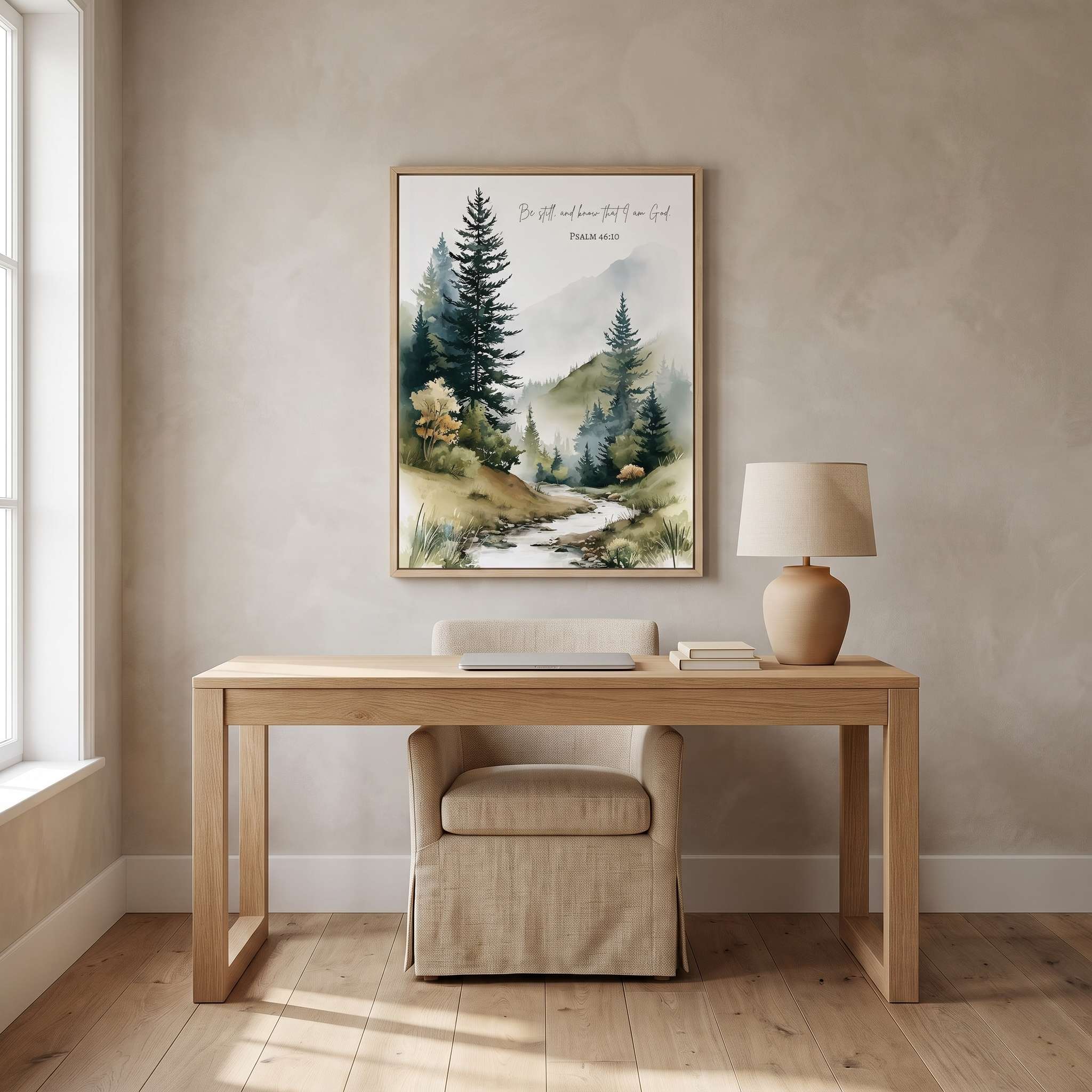 Framed Christian mountain landscape wall art titled Psalm 46:10 featuring evergreen trees, misty mountains, and a peaceful flowing stream, displayed on a beige wall above a wooden desk in a bright modern home office.
