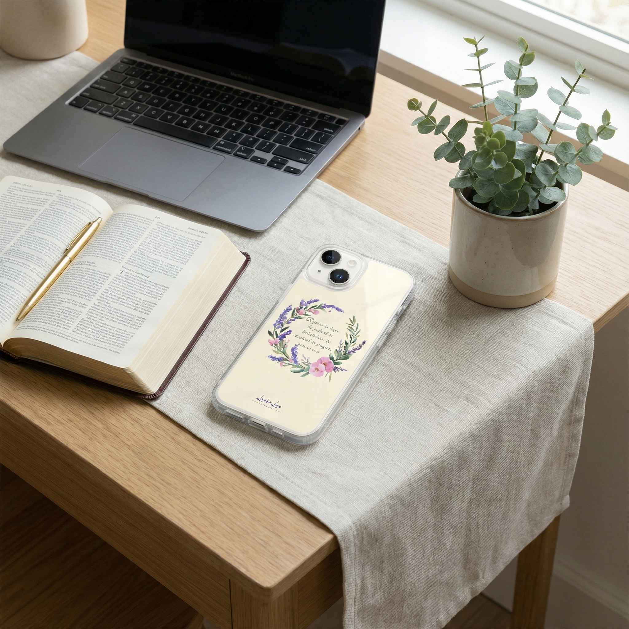 Laptop, open book, phone with floral case, and potted plant on a wooden desk.