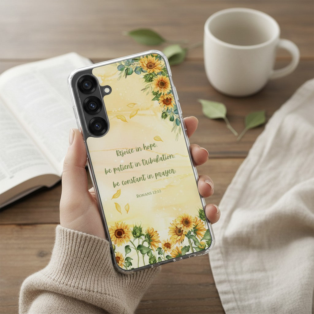 Hand holding a Christian Samsung Galaxy phone case with yellow sunflowers and Romans 12:12 Bible verse, set against a cozy aesthetic background with a white mug and open book.