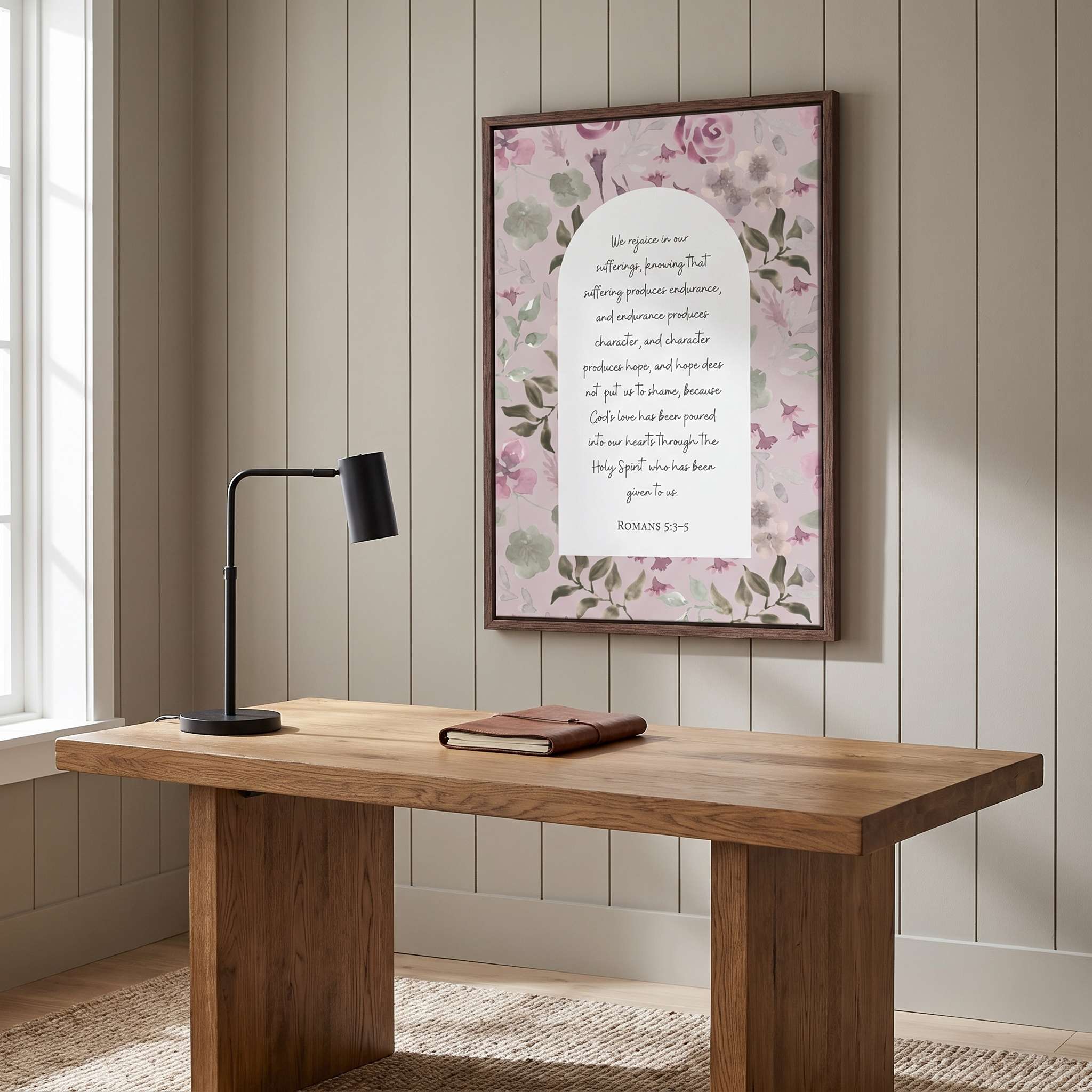 Framed vintage Christian floral wall art titled Romans 5:3–5 featuring "Hope does not put us to shame" with watercolor botanical artwork, displayed in a dark wood frame on a beige paneled wall above a rustic wooden desk with a black lamp in a cozy office.