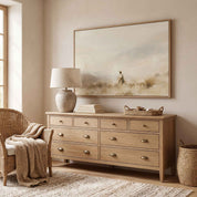 Framed vintage fine art canvas painting of Jesus leading sheep hanging on a warm beige wall above a light natural wood dresser with a ceramic lamp and wicker armchair in a neutral bedroom.