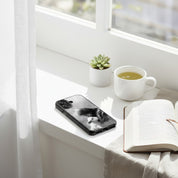 The Good Shepherd MagSafe® clear iPhone case featuring black and white watercolor artwork of Jesus holding a lamb beside an open book, tea mug, and small plant on a bright minimalist windowsill in a peaceful Christian lifestyle setting