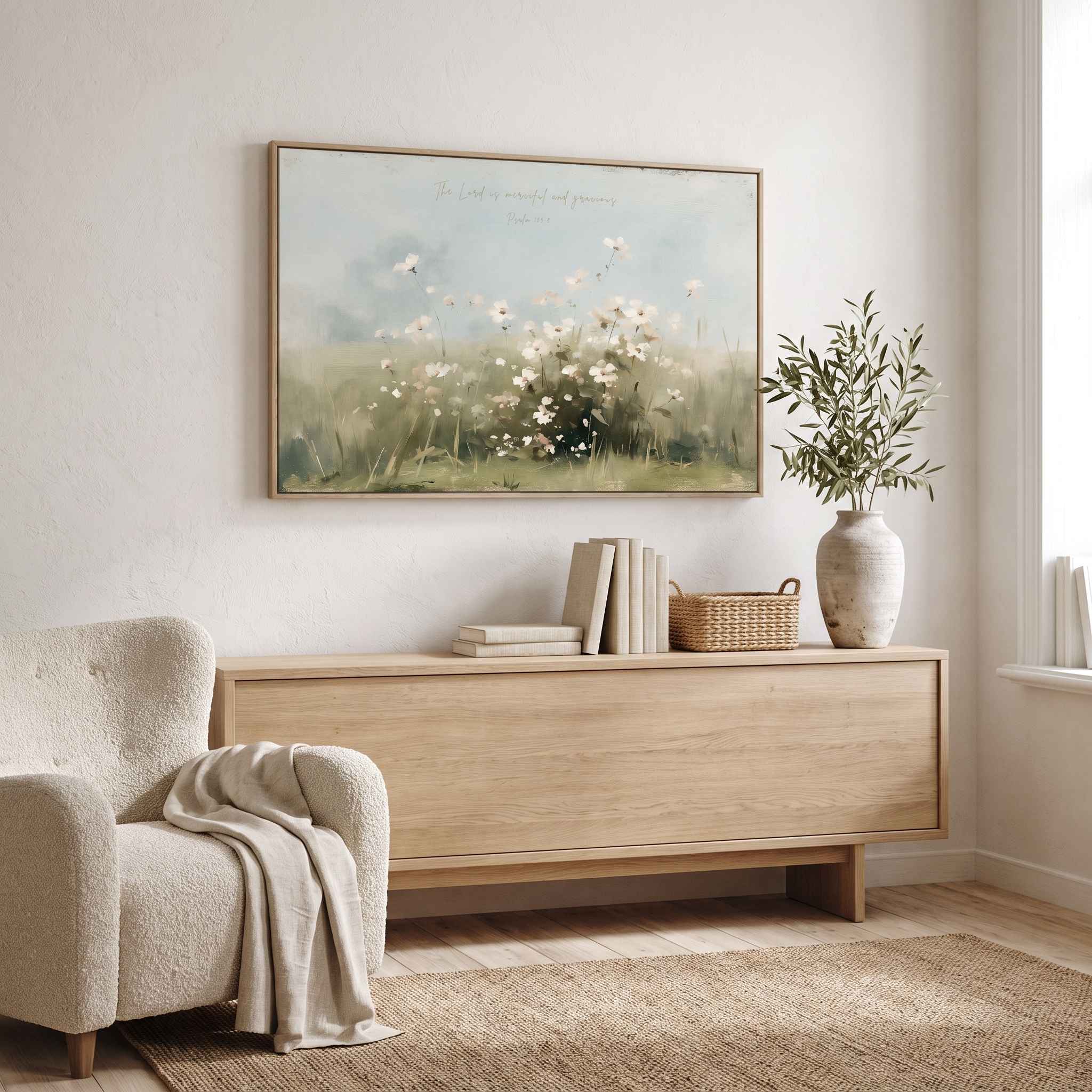 Framed vintage Psalm 103:8 wildflower landscape canvas wall art hanging above a light wood credenza next to a boucle armchair in a bright organic modern living room.
