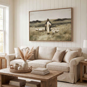 A large framed vintage Christian art print depicting Jesus walking with a flock of sheep, displayed on a white paneled wall above a beige fabric sofa in a bright, modern living room.