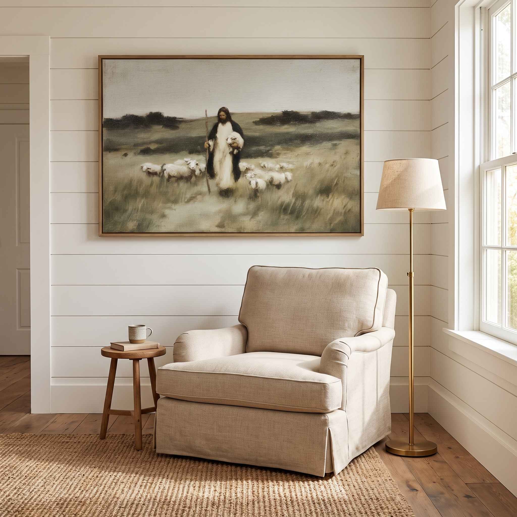A large framed vintage Christian art print depicting Jesus walking with a flock of sheep, displayed on a white shiplap wall above a beige armchair in a bright modern reading nook with a floor lamp.