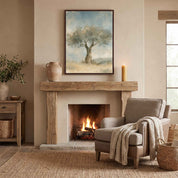 Framed Trust in His Steadfast Love olive tree Christian wall art above a rustic fireplace with a wooden mantel, pottery and candle beside an armchair.