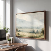 Framed vintage Proverbs 3:5-6 Christian landscape canvas wall art featuring a misty valley scene hanging on a light gray wall above a wooden home office desk with a computer monitor, small plants, and stacked books.