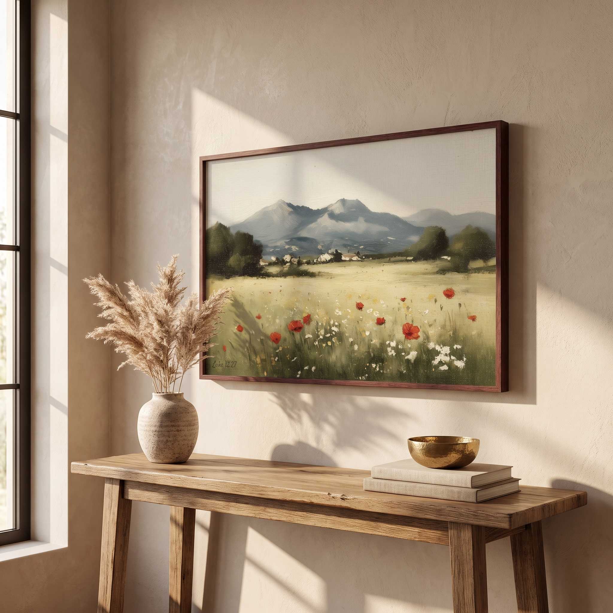 A framed landscape painting of a mountain valley with red poppies hangs on a beige wall above a rustic wooden console table styled with a vase of dried grass and a gold bowl.